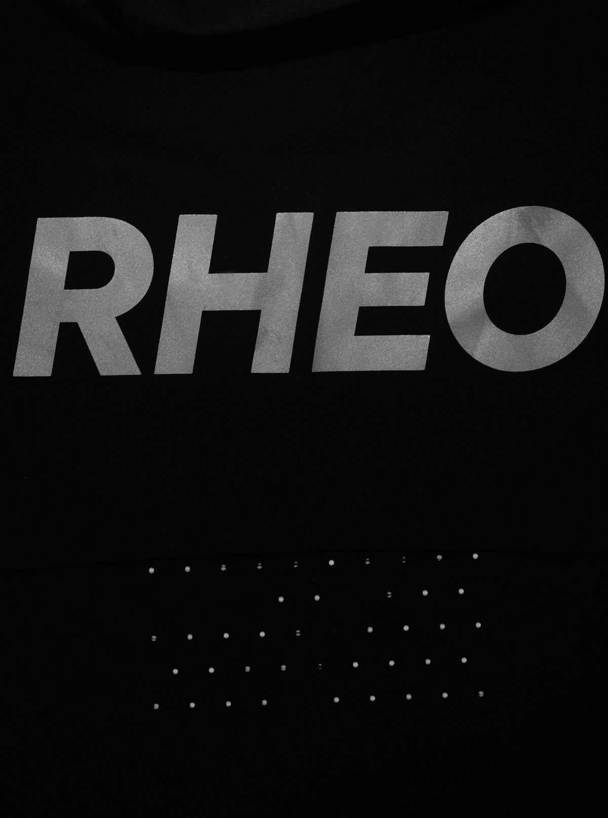 Close-up of a black garment featuring a large metallic silver 'RHEO' logo across the chest and a grid of small silver studs below.