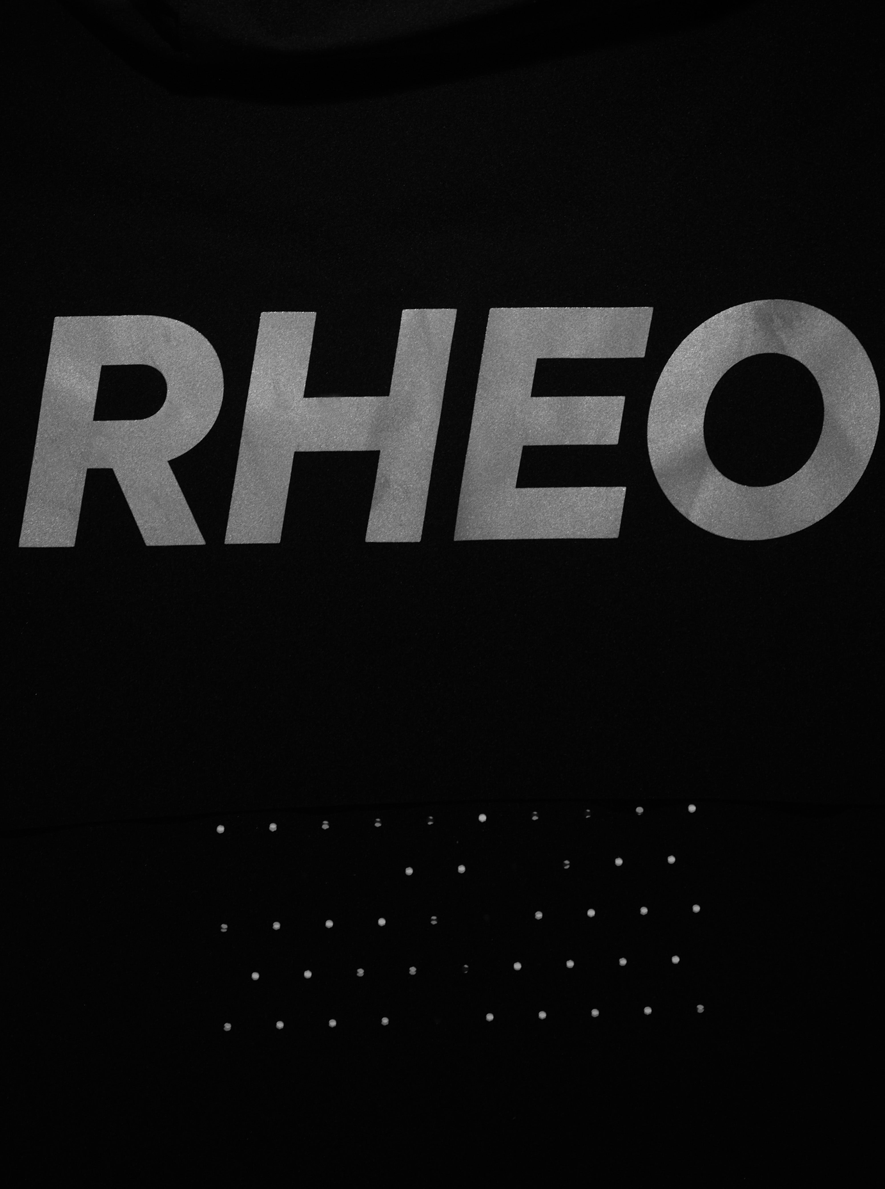 Close-up of a black garment featuring a large metallic silver 'RHEO' logo across the chest and a grid of small silver studs below.