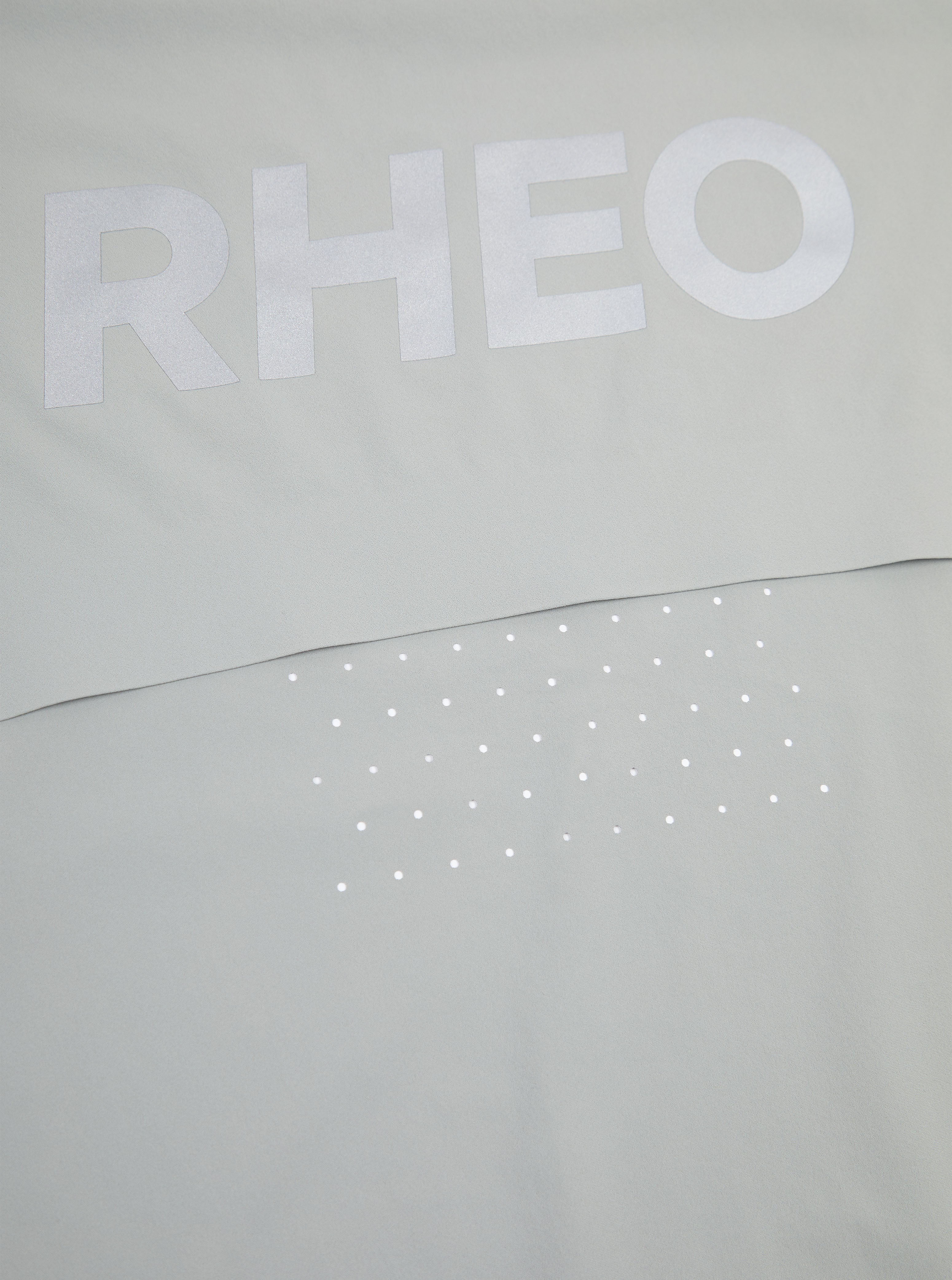 Close-up of a light gray garment with a large white 'RHEO' logo across the chest and a row of small white dot embellishments below.